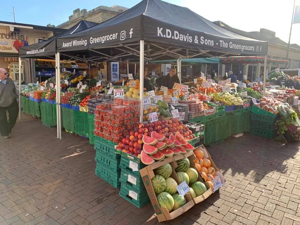 A branded black gazebo being used as shelter for a fruit and veg stall