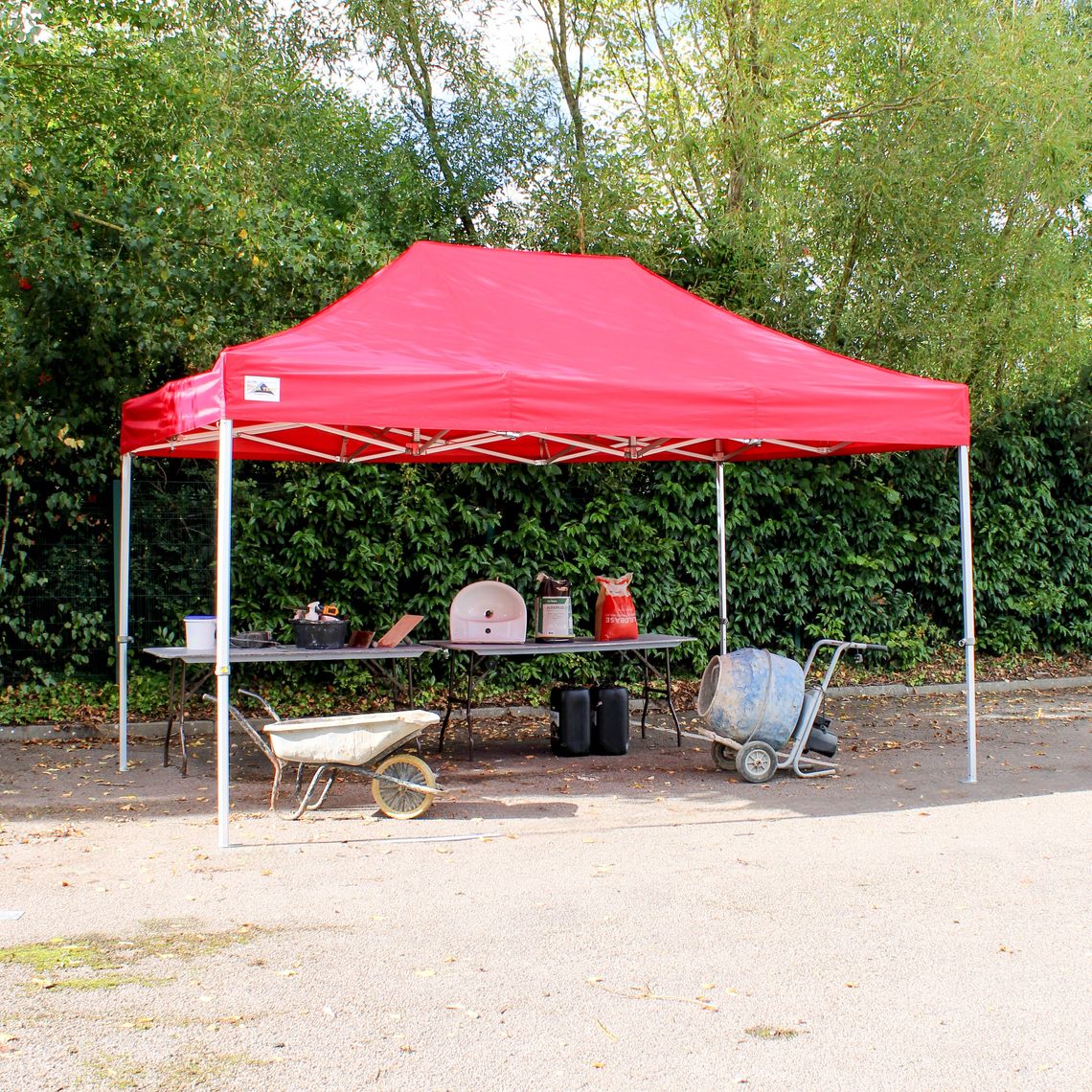 A red pop up gazebo with work tools beneath the canopy, in front of green trees